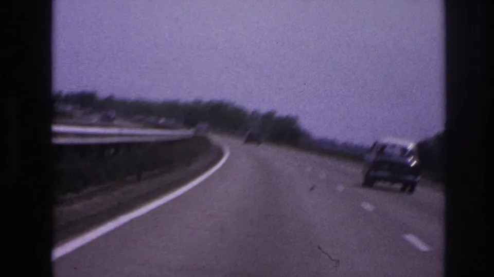 Forward 8mm view of a 1967 Paris-area highway, with a car ahead on the curve and roadside trees beyond a white guard rail.