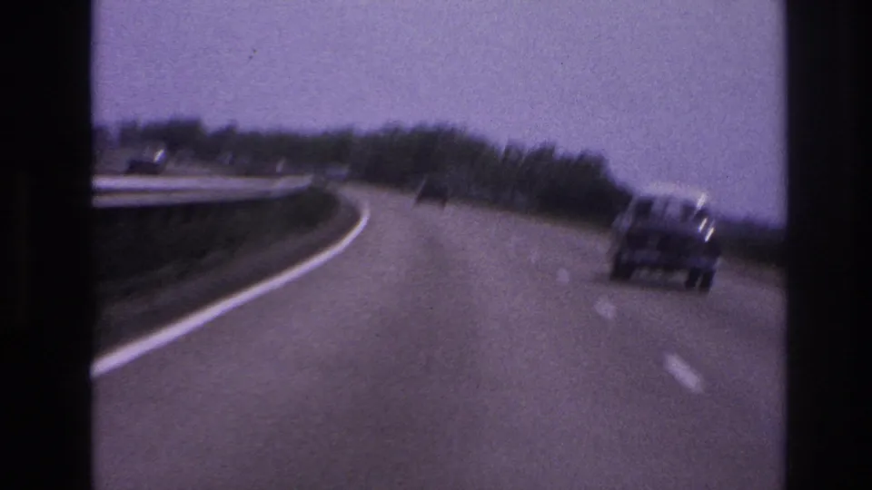1967 8mm frame from Paris, France metadata showing a curved paved highway with a car ahead, roadside trees, and guardrail edges.