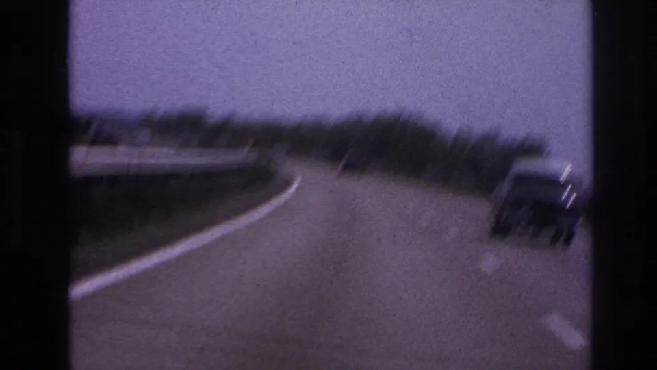 1967 8mm frame from the Paris, France clip showing a vehicle on a curved highway with roadside guardrails, trees, and soft motion blur.