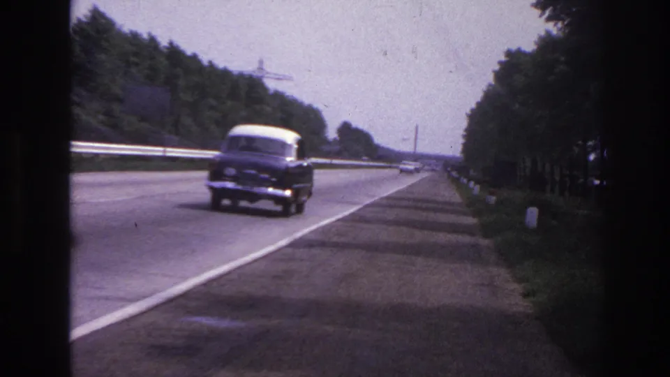 1967 8mm frame from Paris, France metadata showing a classic car approaching on a paved roadway with shoulder, trees, and guardrails.