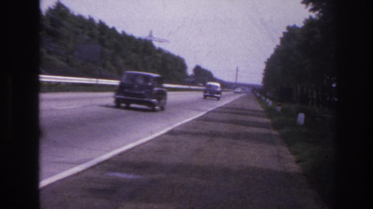Dark sedan and smaller cars driving along a tree-lined highway with a guardrail.