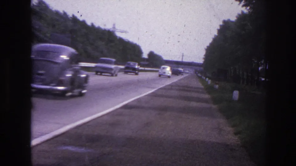1967 Paris-area highway frame with a dark car blurred in the foreground and several vehicles ahead on the tree-lined road.