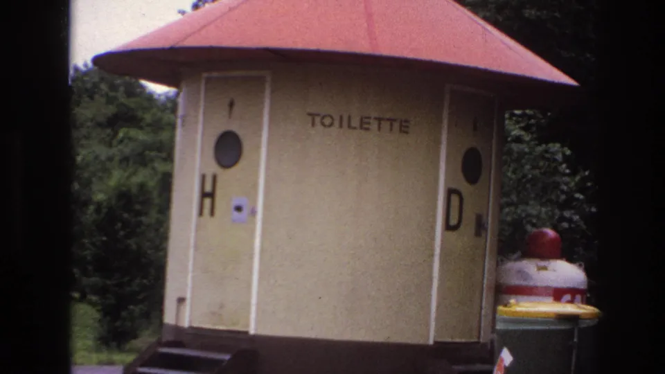 Final 1967 home-movie frame holding on the roadside TOILETTE kiosk, red roof, door markings, trees, and nearby red-topped fixture.