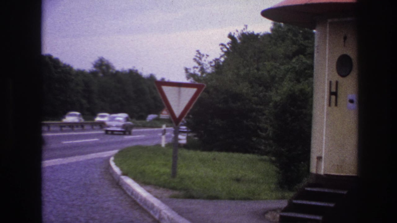 1967 Paris-area 8mm frame showing cars on a multi-lane road, a red-bordered yield sign, grassy verge, and a small roadside structure.