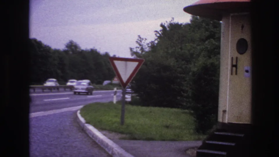 1967 Paris-area 8mm frame showing cars on a multi-lane road, a red-bordered yield sign, grassy verge, and a small roadside structure.