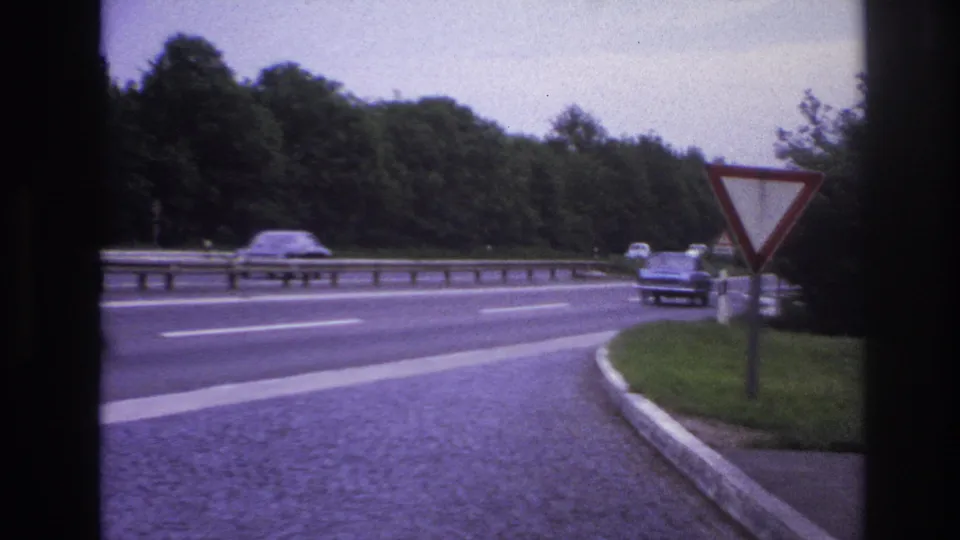 1967 Paris-area 8mm still showing an approaching car near a red-bordered yield sign, metal guardrail, cobbled shoulder, and dense roadside trees.