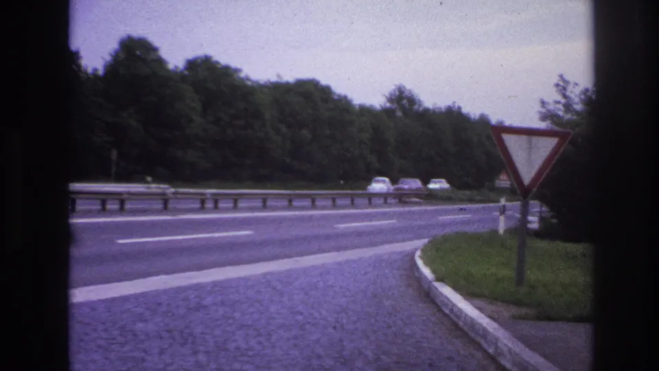 1967 Paris-area road scene with a red-bordered yield sign, distant cars, metal guardrail, grassy verge, cobbled shoulder, and wooded background.