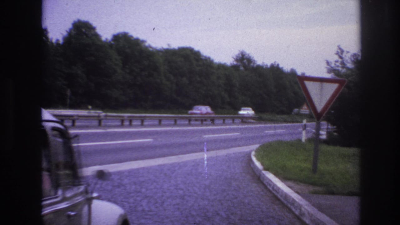 Paris-area 1967 home-movie frame showing a roadside merge, yield sign, guardrail, trees, and a partial car from inside a vehicle.