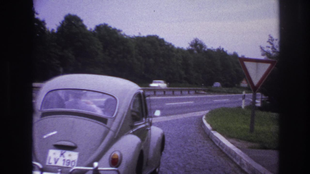 1967 Paris-area frame of a grey VW Beetle with visible rear plate approaching a yield sign near highway lanes and trees.