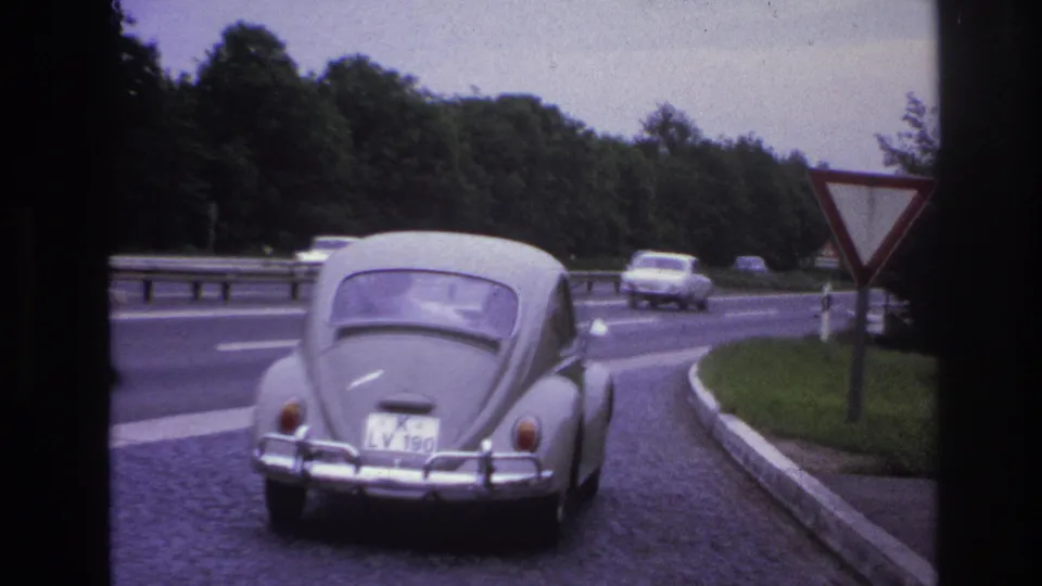 Paris-area 1967 home-movie frame showing the rear of a grey VW Beetle on a cobblestone merge beside highway traffic.