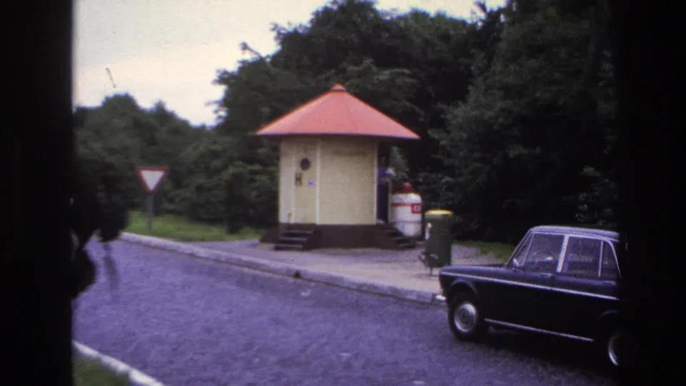 1967 Paris-area roadside frame with a yellow kiosk, red roof, cobblestone road, trees, traffic signs, and a dark period car.
