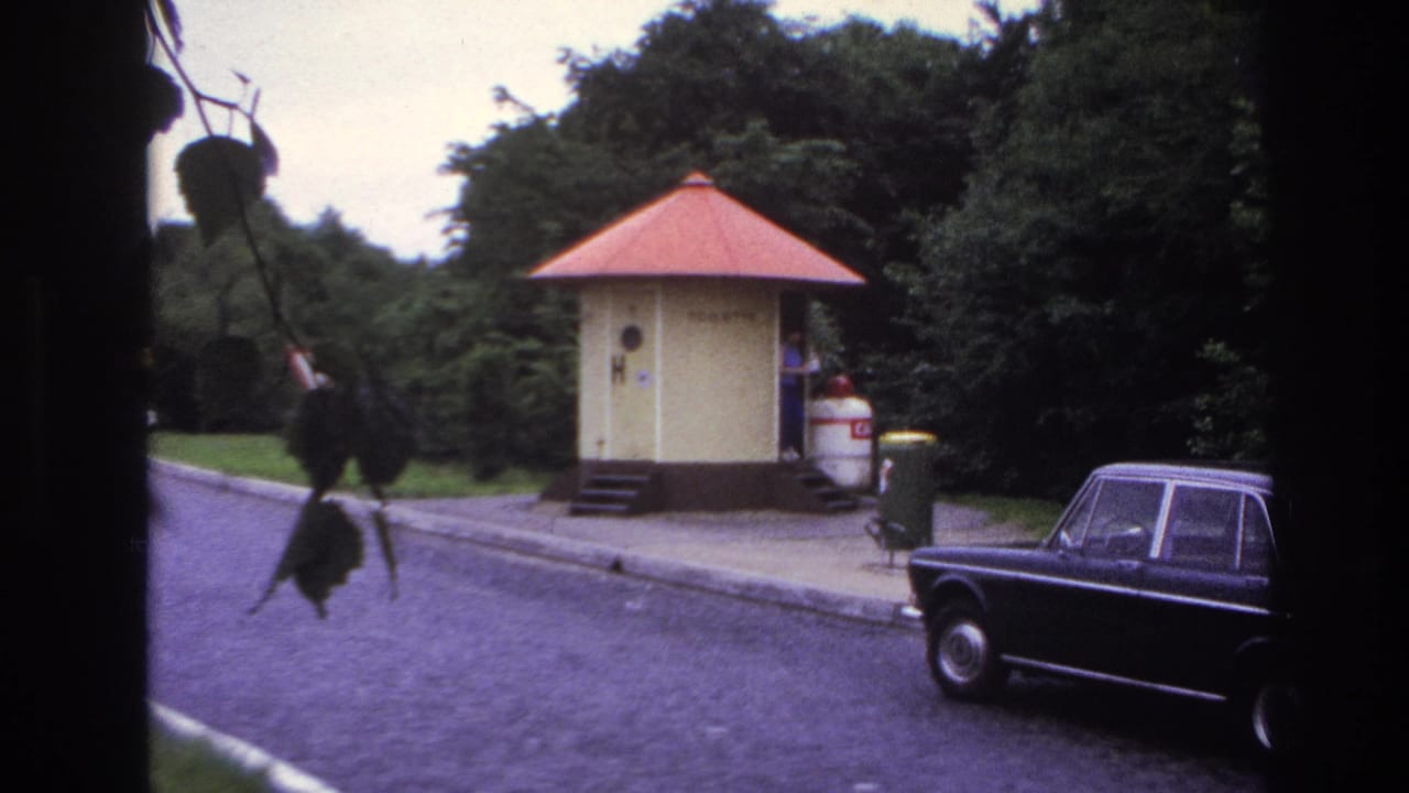 1967 Paris home-movie frame showing a red-roofed yellow kiosk, parked dark sedan, curb, trees, and foreground leaves.