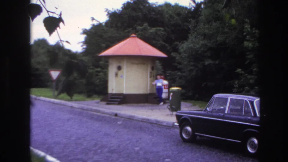 1967 Paris home-movie view of a woman leaving a small yellow kiosk, with a dark sedan, bins, trees, and curbside pavement.