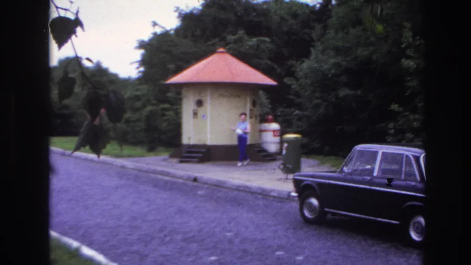1967 Paris frame showing a woman outside the red-roofed kiosk, with a dark sedan parked along the curb and leafy trees around the scene.