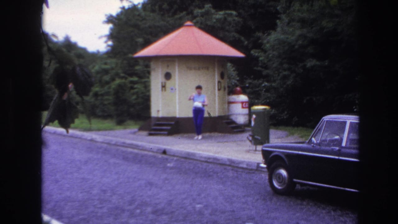 1967 Paris home-movie frame showing a woman near yellow public toilet kiosks, a curbside road, greenery, and a dark parked car.