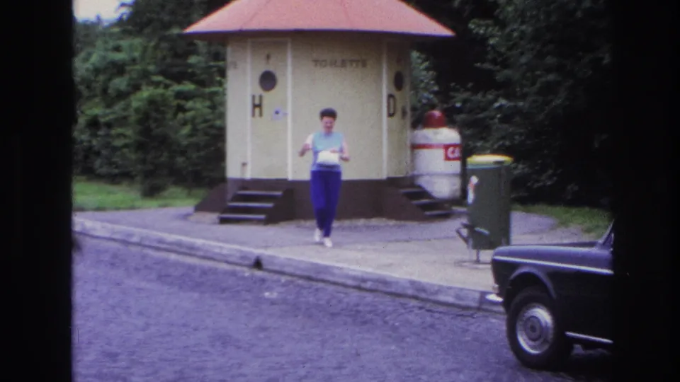 1967 Paris frame with a woman carrying a light bag near public toilet kiosks, trash bins, a curb, and a dark car at frame edge.