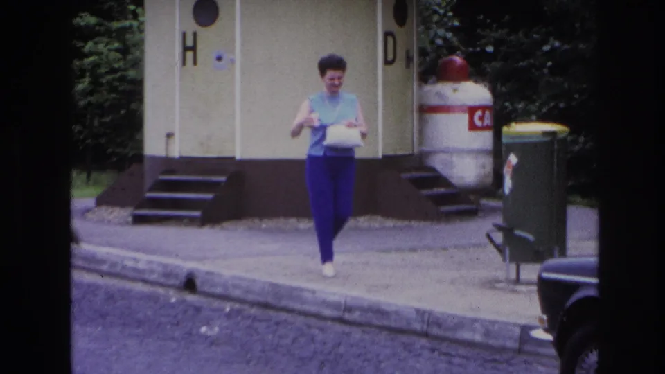 Closer 1967 Paris home-movie view of a woman in blue and purple walking past yellow public toilet doors with bins and foliage nearby.