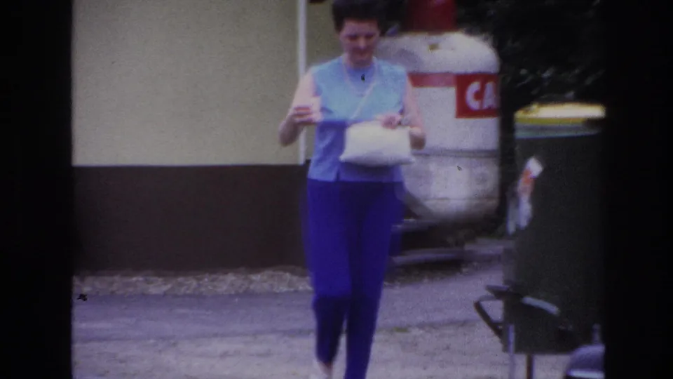 Close 1967 Paris frame of a woman walking with a white bag, with part of the public toilet structure, signage, bins, and road edge visible.