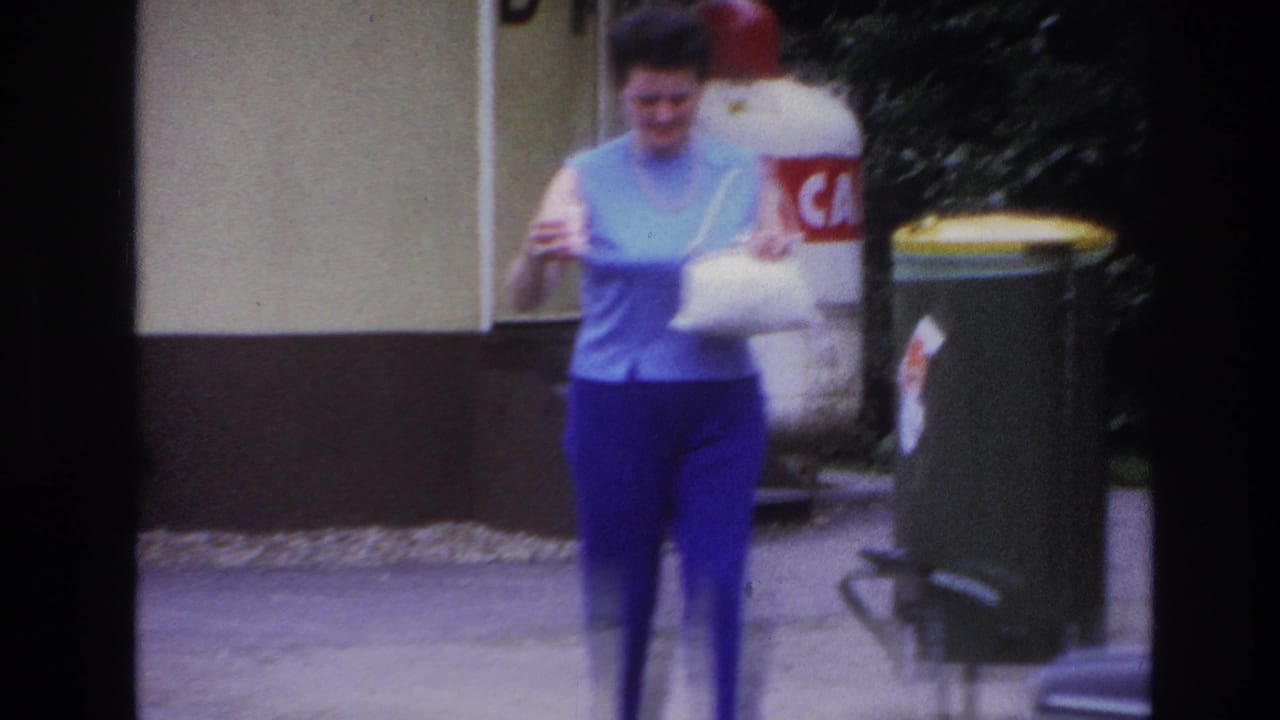 In 1967 Paris home-movie footage, a woman in a light blue sleeveless top walks on a sidewalk carrying a white bag and cup.
