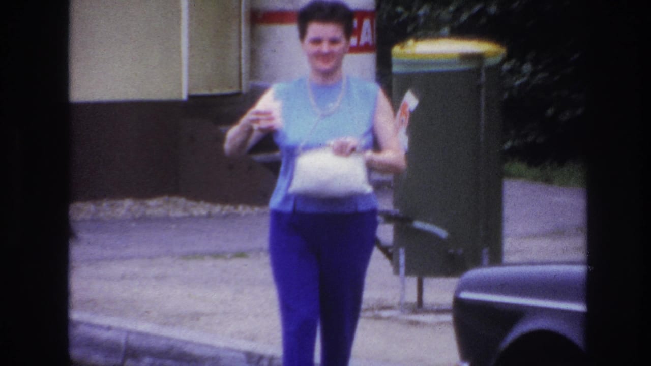 A smiling woman walks toward the camera on a Paris sidewalk in 1967, holding a white bag and small cup near a curbside bin.