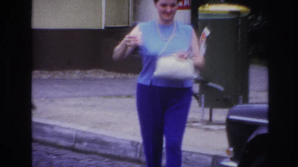 Closer 1967 Paris 8mm view of the woman walking toward the camera with a white bag, cup, curb, bin, and car edge visible.
