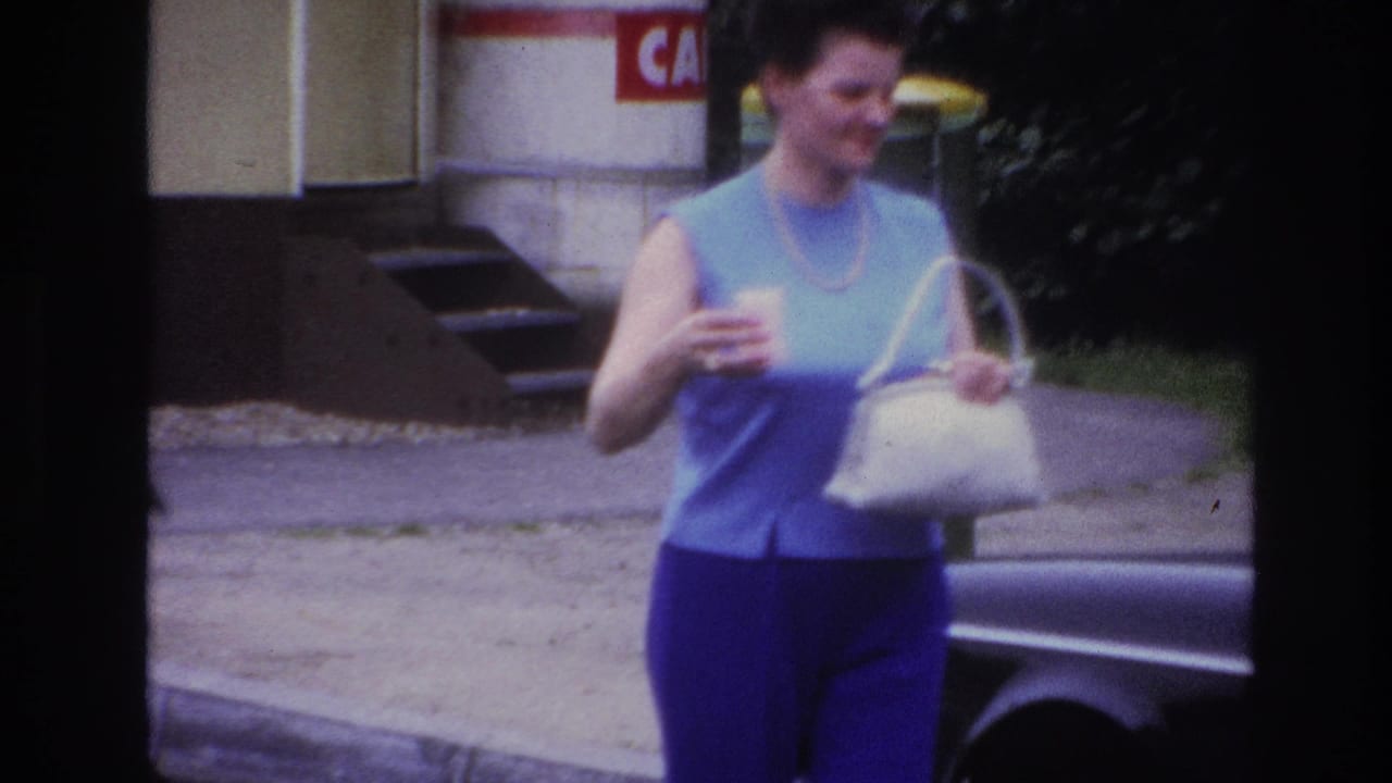 In 1967 Paris home-movie footage, a woman approaches a dark car while carrying a white handbag and small cup.