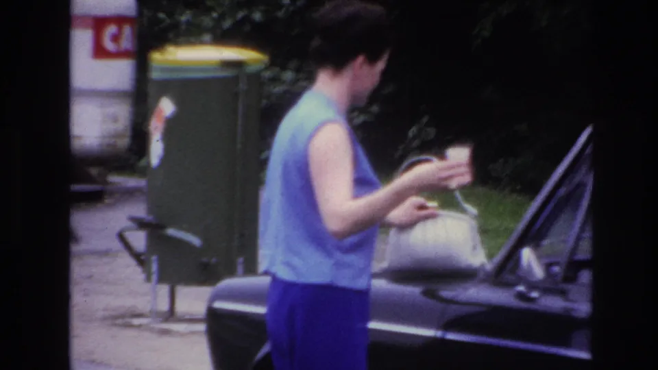 The woman stands at the side of a dark car in Paris, holding a cup while positioning her white handbag near the hood.