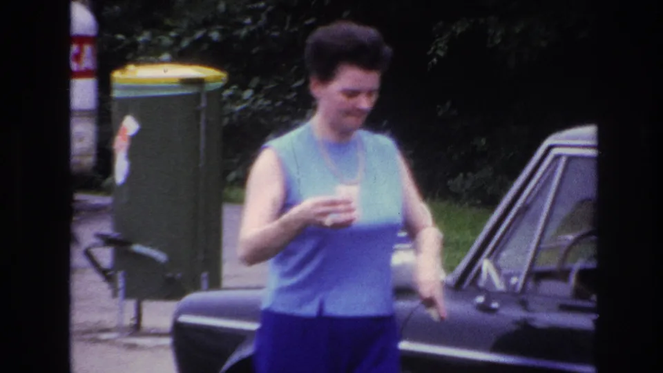 The woman smiles beside the dark car in the 1967 Paris home movie, with her handbag lowered and a cup still in hand.