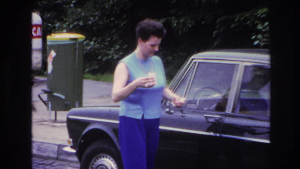 In 1967 Paris home-movie footage, a woman stands by a dark sedan, holding a small cup near the front door.