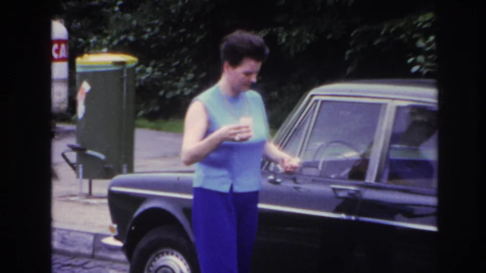 In 1967 Paris home-movie footage, a woman stands by a dark sedan, holding a small cup near the front door.