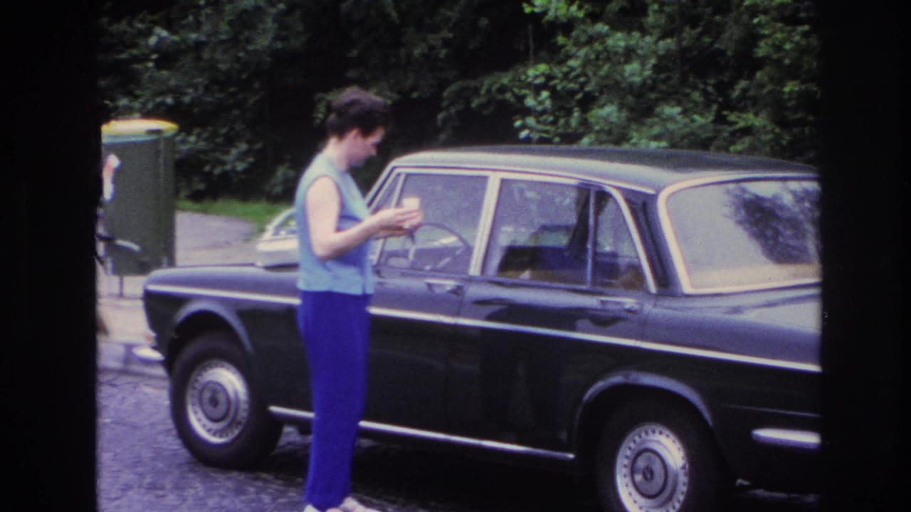 Woman stands near the front door of a dark sedan with trees behind the street.