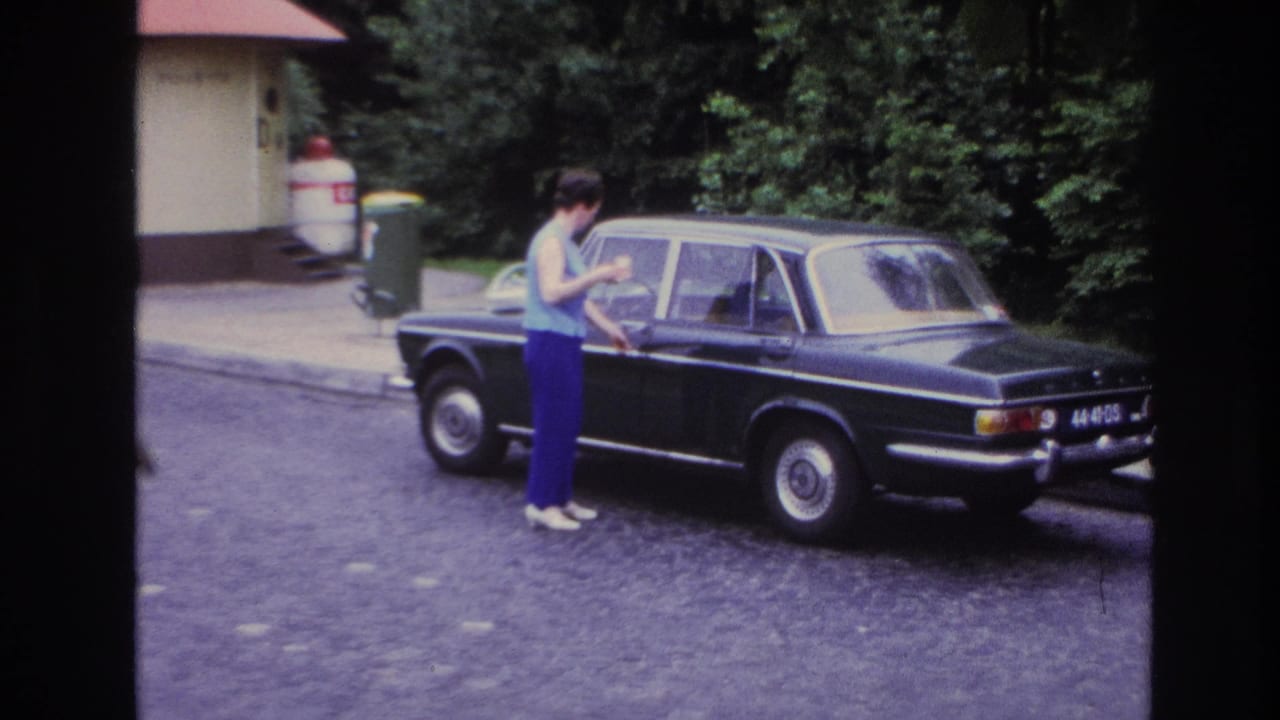 In 1967 Paris home-movie footage, a woman stands by the rear door of a black sedan on a cobblestone street.