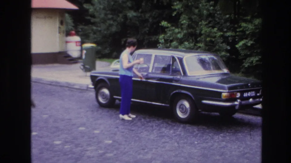 In 1967 Paris home-movie footage, a woman stands by the rear door of a black sedan on a cobblestone street.