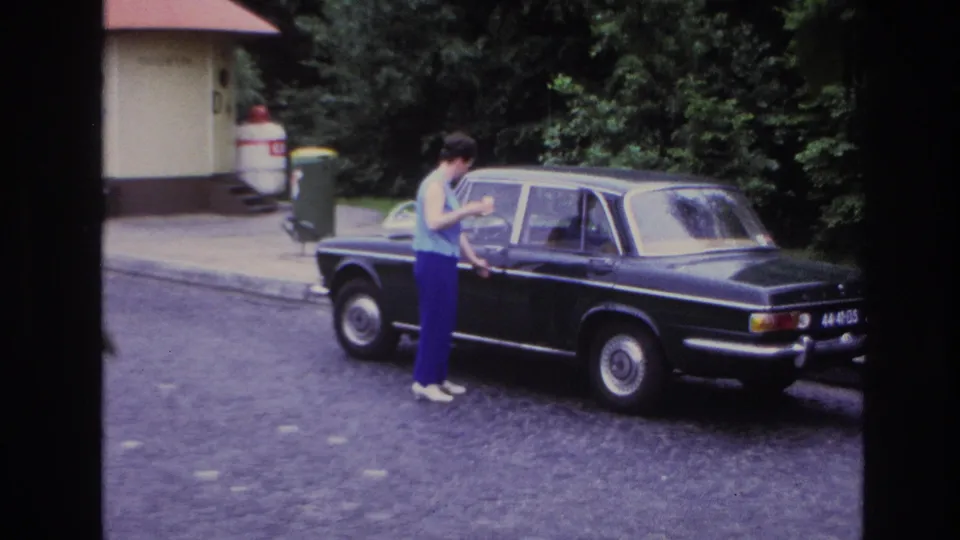 A woman in a light top and purple trousers reaches for a black sedan door in 1967 Paris footage.
