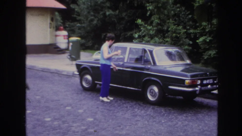 The woman stays beside the black sedan, framed by trees and a small building in Paris home-movie footage.