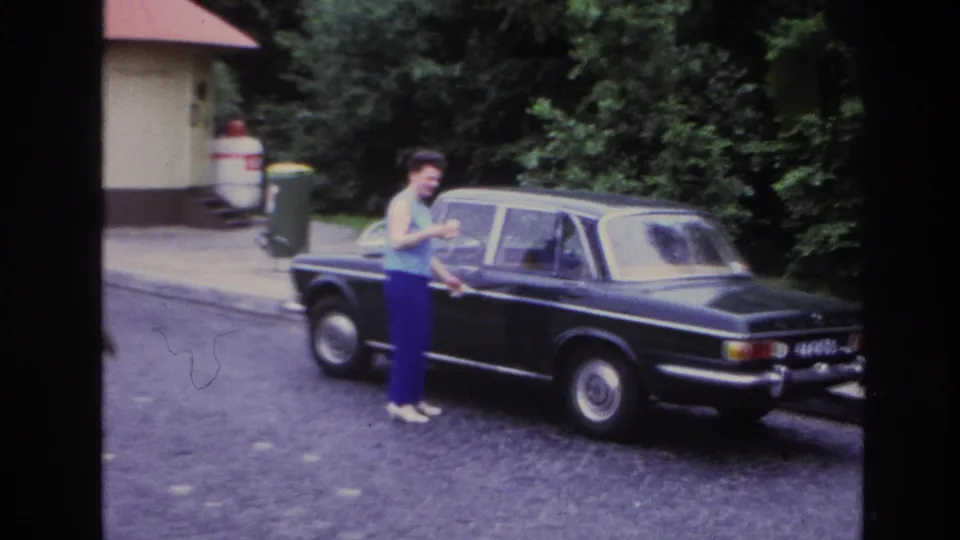 A woman glances toward the camera beside a black sedan on a Paris street in 1967 home-movie footage.