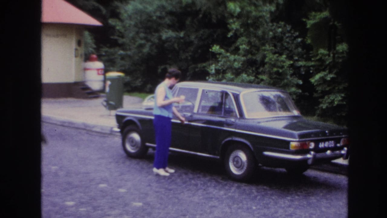 1967 Paris home-movie frame showing a woman unlocking or opening a black sedan on a cobblestone street beside trees and a small red-roofed building.