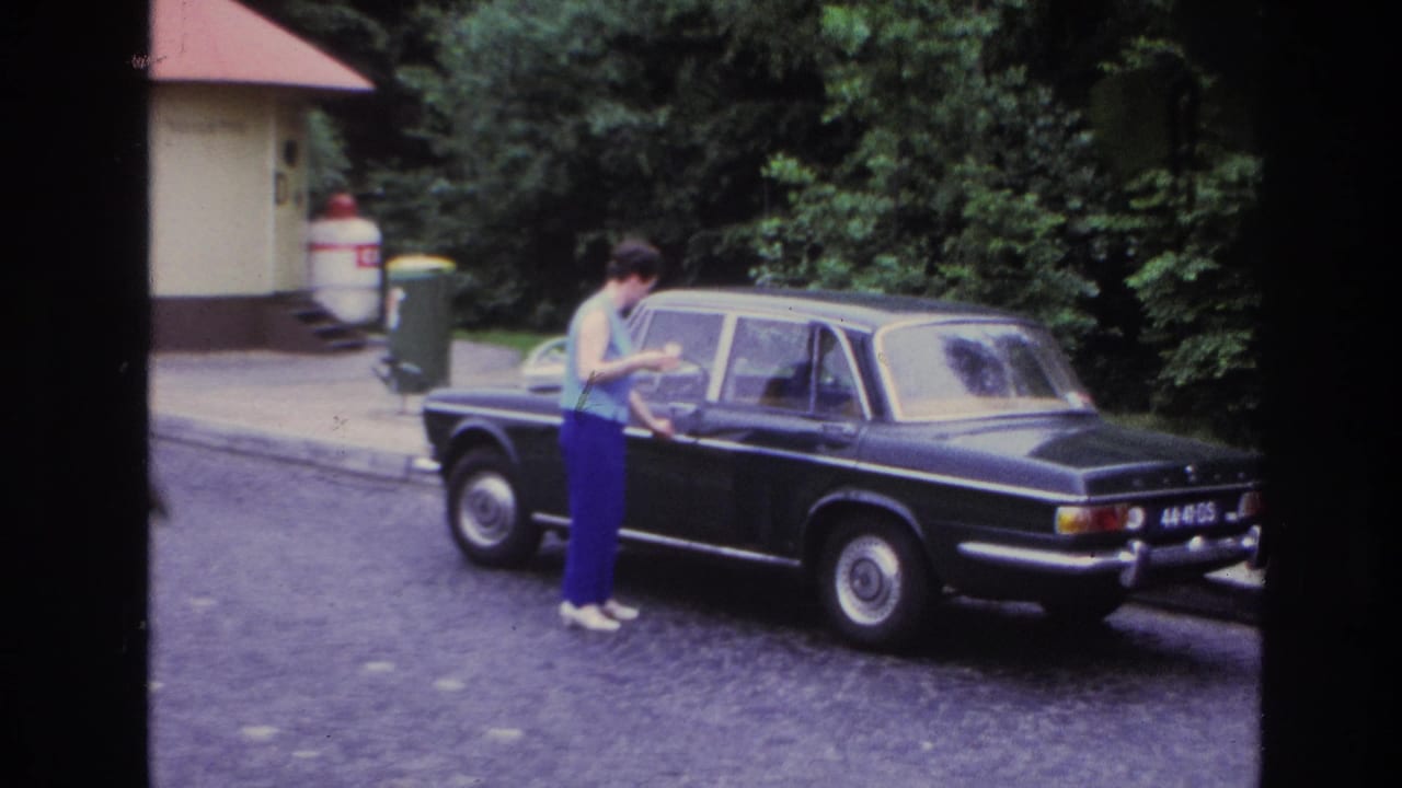Woman beside the same black sedan, with trees and a small building in the background.