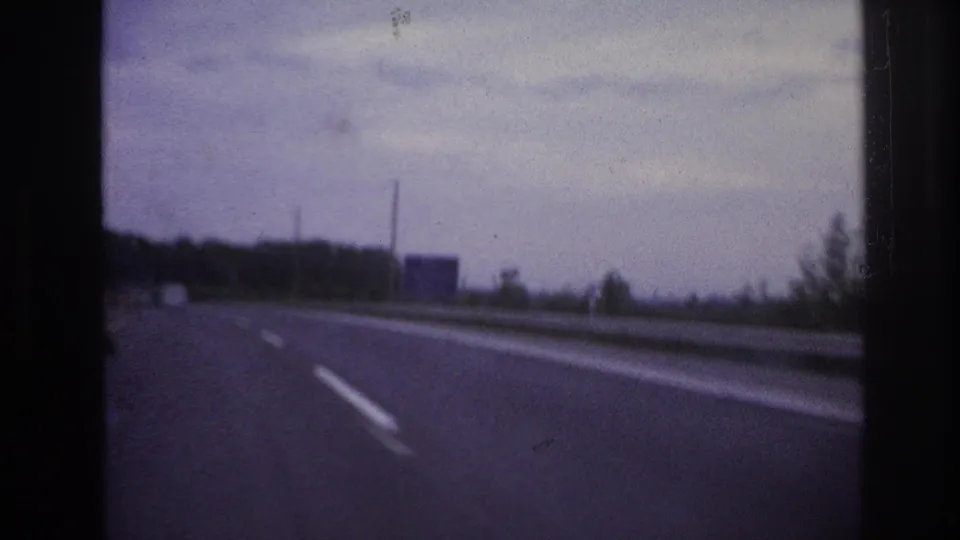 Home-movie road view after the car scene, showing a highway lane, guardrail, trees, poles, and a distant roadside sign under a cloudy sky.