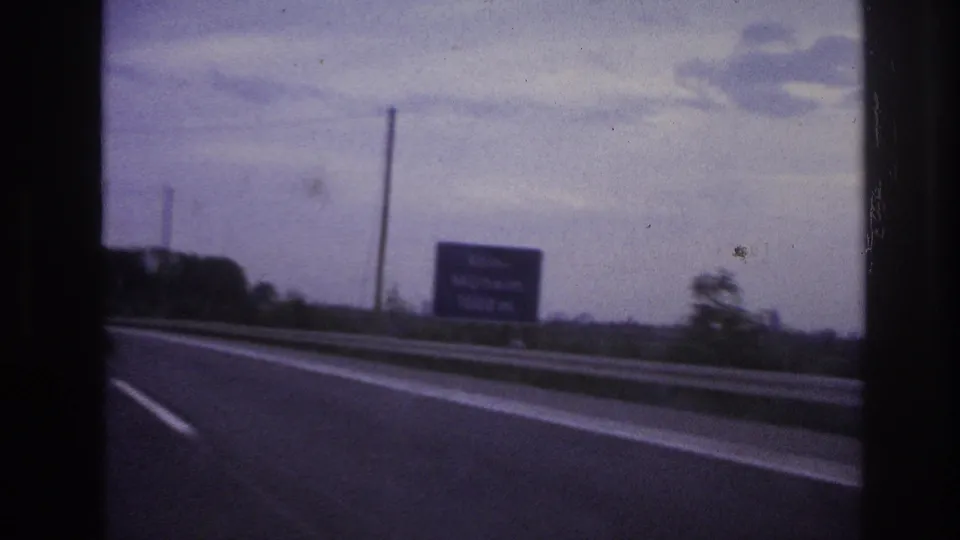 Home-movie frame of a highway or major road, with a roadside sign, guardrail, dark tree line, and muted cloudy sky visible through the film frame.