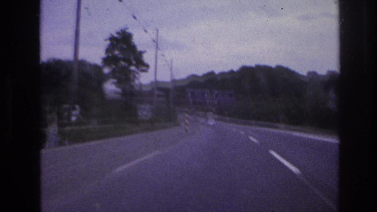 1960s 8mm home-movie view from inside a moving vehicle on a European highway with trees, lane markings, and overcast sky.