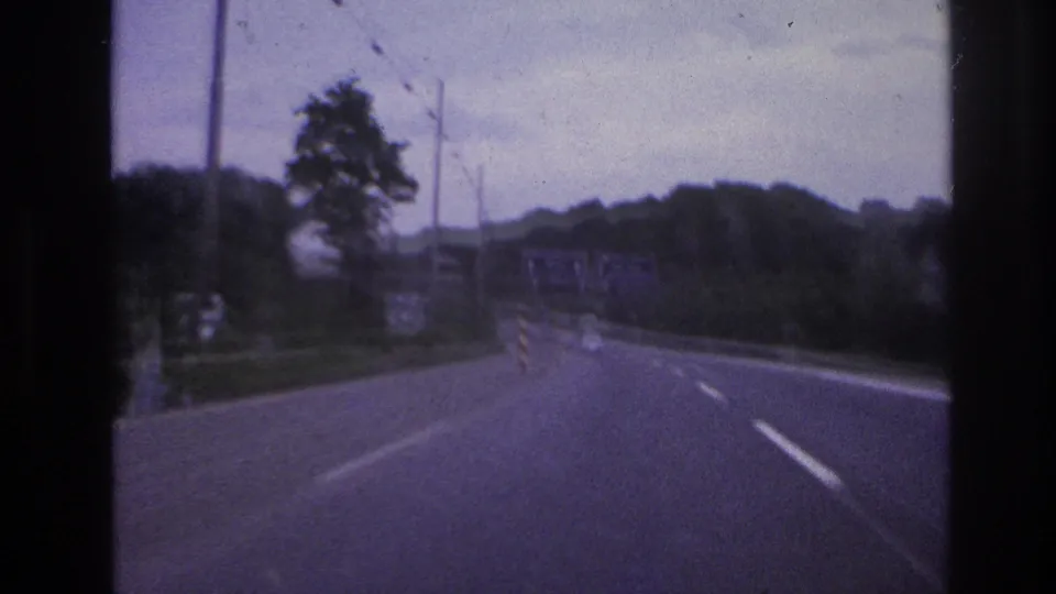 1960s 8mm home-movie view from inside a moving vehicle on a European highway with trees, lane markings, and overcast sky.