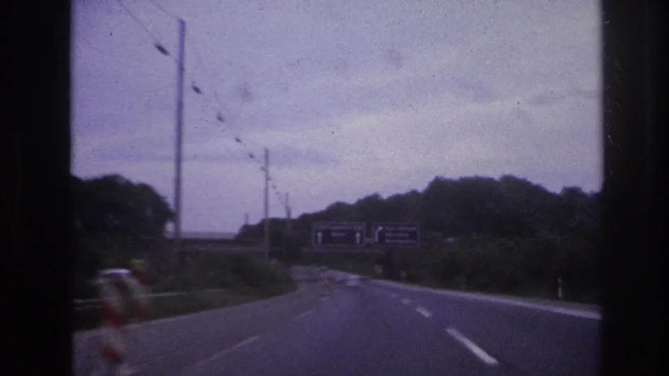 Moving-vehicle view on a highway approaching overhead direction signs, with wooded roadsides and a cloudy sky visible.