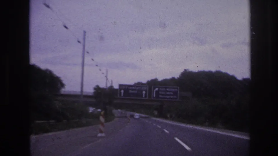European highway view with overhead signs for Frankfurt and Bonn partly legible, filmed from a moving vehicle in 8mm.
