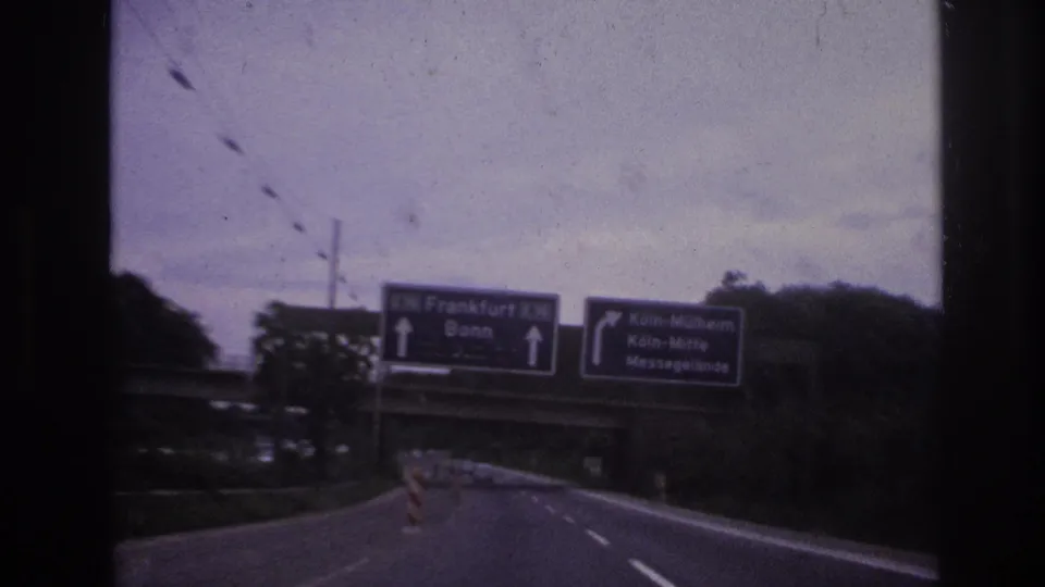 Close moving-vehicle view of German highway direction signs for Frankfurt, Bonn, and Köln-Mülheim, with trees and an overpass nearby.