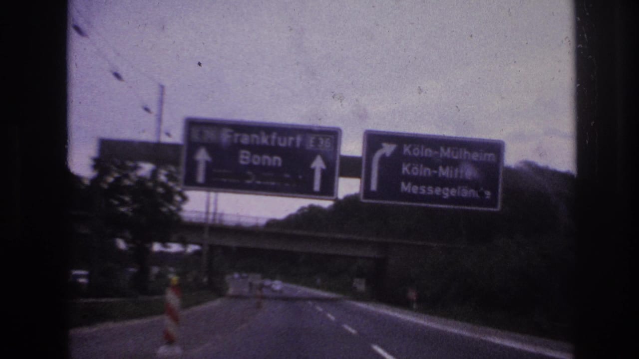 1960s 8mm road view from inside a moving car, with German motorway direction signs above a divided roadway.