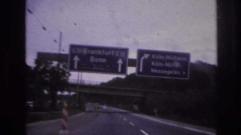 1960s 8mm road view from inside a moving car, with German motorway direction signs above a divided roadway.
