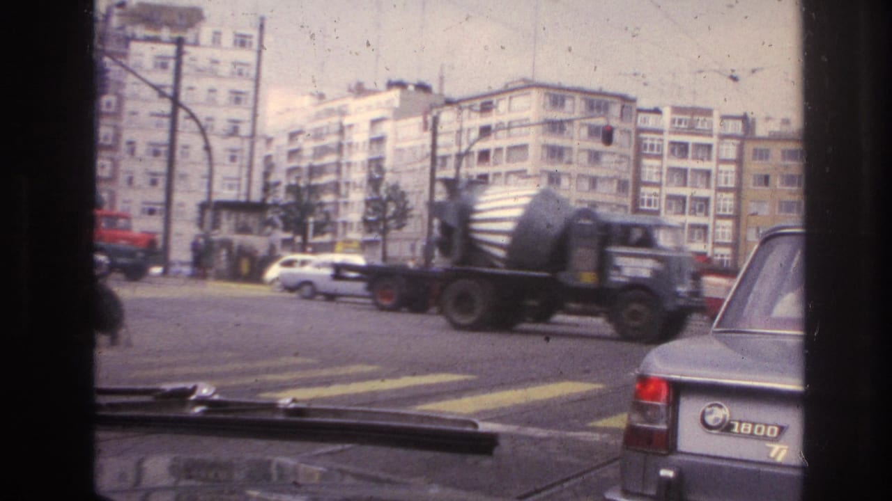 Urban intersection with a red cement mixer truck, crosswalk markings, apartment blocks, and a BMW 1800 sedan.