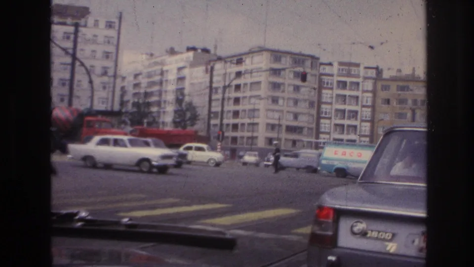 Street traffic in the claimed 1967 travel footage, with compact cars, a blue van, overhead wires, and multi-story residential buildings.