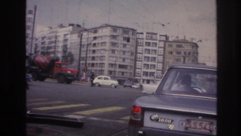 Late-1960s city street view from a car, showing a cement mixer, compact vehicles, overhead wires, and apartment buildings.
